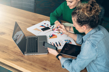 Teamwork. Two young business women sitting at table in front of laptop. On table is tablet computer and paper charts. First woman looking at screen of smartphone, other takes notes in notebook.