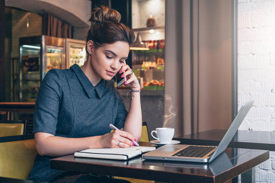 Young business woman in gray dress sitting at table in cafe, talking oncell phone while taking notes in notebook. On table laptop and cup of coffee. Student learning online. Freelancer working online.