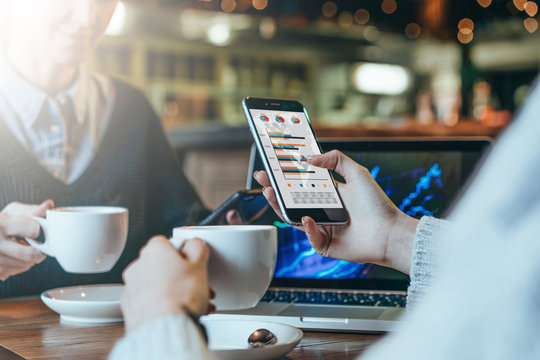 Close-up of a smartphone in female's hand. Graphs and charts on smartphone screen.Two young business women sitting at table,drinking coffee and analyzing data.On table laptop.Students studying online.