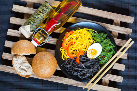 Hiyashi wakame with sesame and nut sauce, black and corn noodles, boiled egg. Bottle of olive oil with chili pepper, black olives and capers in jar, bran bread. The rustic wooden lattice, black makisu