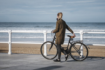 A blonde girl walks with her bike in her hand for a walk on the beach of a city