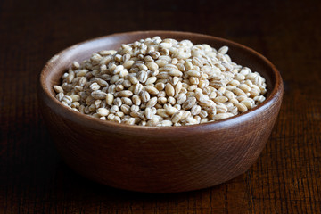 Dry pearl barley in brown wooden bowl isolated on dark wood.