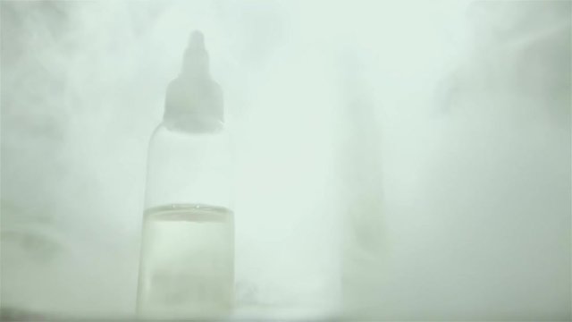 Bottle with liquid and flavor for electonic cigarette stay on the table. A lot of steam for background.