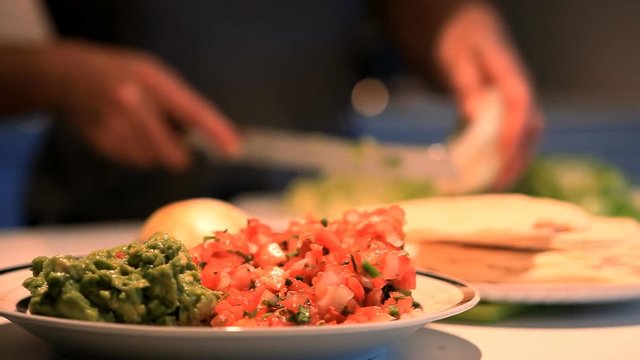 Focus on salsa and guacamole with a person out of focus in the background  preparing a Mexican meal.