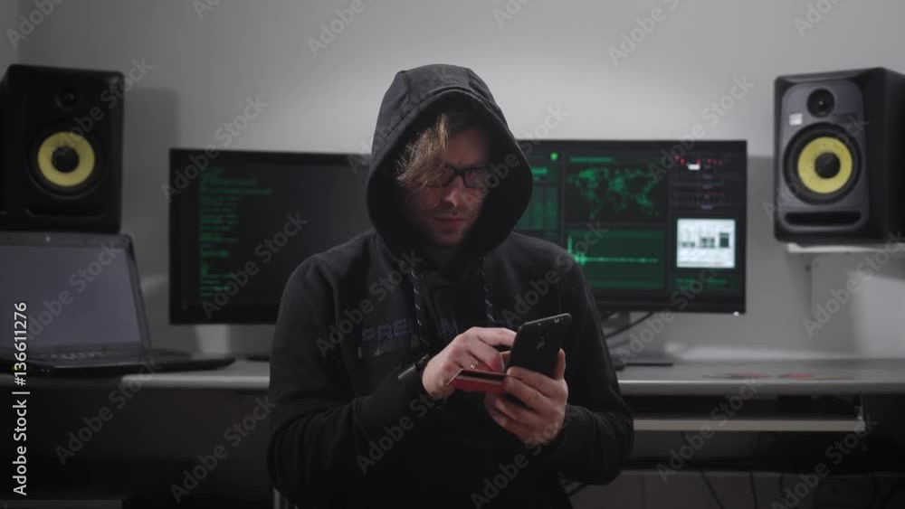 Poster close up of a hooded hacker making cyber attack on bank network with the help of smartphone in his h