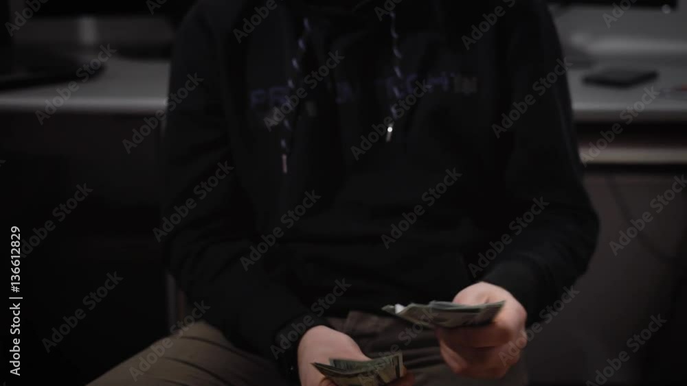 Poster close up of a hooded hacker with eyeglasses dressed in black clothes sitting on a chair in the room 