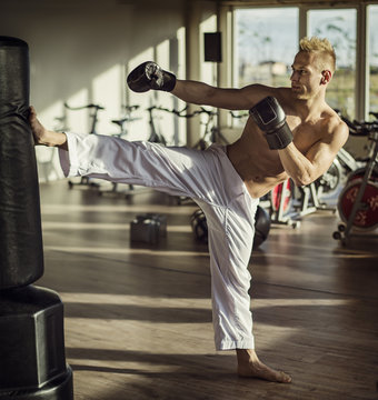 Shirtless handsome muscular young man in gym doing kick against punching bag, wearing boxing gloves