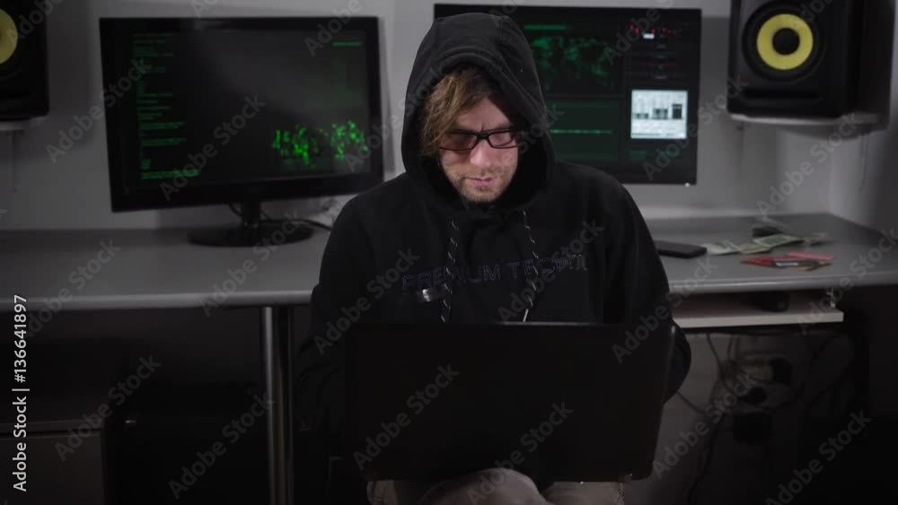 Poster young hacker in black working on laptop sitting alone in the room with computer monitors behind him.