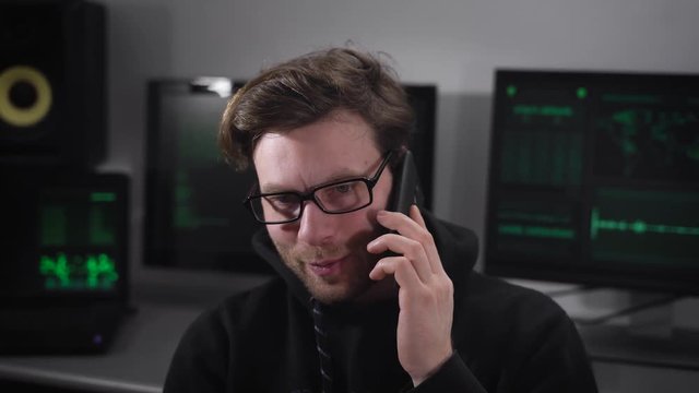 Man with eyeglasses dressed in black clothes is sitting in the room and speaking on the telephone. Young hacker is coming to an agreement with the customer about money for the future cyberattack.