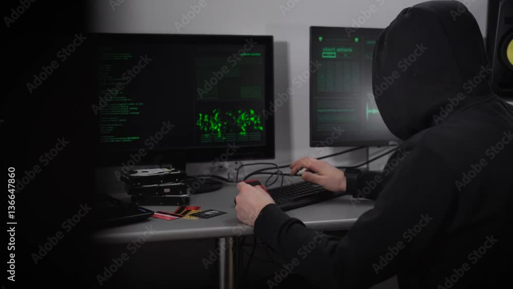 Poster back view of a hooded hacker making cyber attack on bank network. man in black is typing fast on key