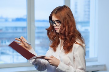 red-haired woman holding a notebook and a pen near the window