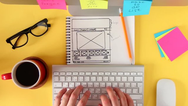 Top view of working place in the yellow table. The web master is working on the computer, making the site layout drawn on the sketch.