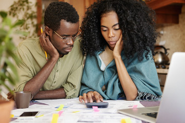 Stressed young African couple can't stand tension of financial crisis, looking unhappy and frustrated, sitting at kitchen table with calculator, trying to save some money by cutting domestic expenses