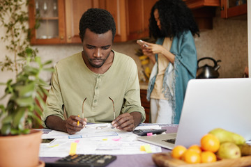 African man with glasses and pencil in his hands looking in frustration at papers in front of him while doing paperwork, trying to pay out all family debts, sitting at table with laptop and calculator