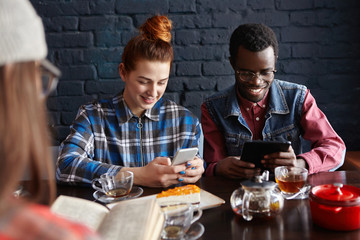 People, communication and modern technology. Internet addicted interracial couple holding their gadgets and using free wi-fi at cafe during meeting with unrecognizable girl with book in foreground