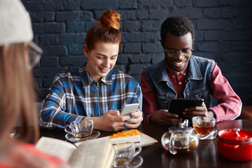 People, communication and modern technology. Internet addicted interracial couple holding their gadgets and using free wi-fi at cafe during meeting with unrecognizable girl with book in foreground