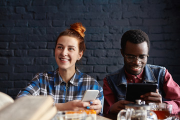 Cute student girl with ginger hair messaging via social networks, her handsome African groupmate sitting next to her with digital tablet, both having cheerful looks, enjoying online communication