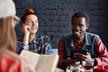 People, technology and communication. Group of three young people having conversation at cafe: redhead woman talking on cell phone, African man using electronic tablet, girl in foreground reading book