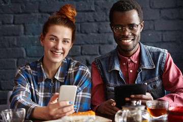Young modern people with electronic devices having fun during coffee break indoors at cafe. Redhead girl texting messages online on her cell phone, her African boyfriend next to her using touch pad