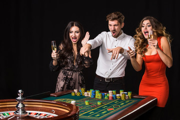 Two young women and man behind roulette table on black background