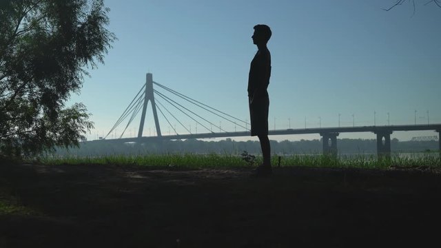 A guy is doing part of his workout. He is stretching his spine bending down and forward. He is training outside in the prk not far from a big town bridge. 