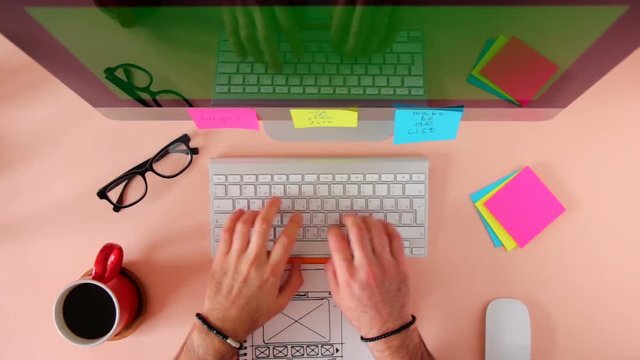 Top view of working place in the pink table. The web master is working on the computer, making the site layout drawn on the sketch.