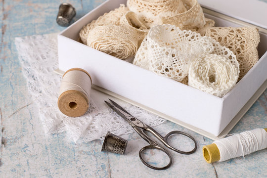  Accessories for needlework. Box with lacy ribbons, spools of thread, thimble and scissors on the old wooden table.
