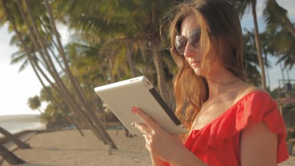 Young woman in a red dress walking by the sea. She is using a tablet and smiling - Powered by Adobe