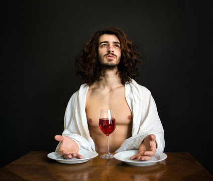 photo half naked handsome guy with long curly hair at the table with two white empty plates and a glass of red wine is praying