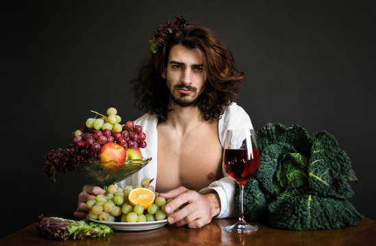 photo half naked handsome guy with long curly hair with a glass of red drink and a plate of grapes and fruits at the table
