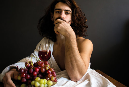 photo half naked handsome guy with long curly hair with a glass of red drink and a plate of grapes at the table