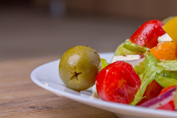 Fresh greek salad in white glass dish plate, closeup from side. Olives, tomatoes, paprika, bell pepper, cucumber, onion, feta cheese, olive oil. Standing at wooden background macro. Vegeterian food.