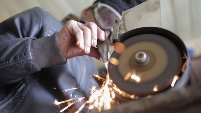 Old man sharpening his ax on a grinder in an workshop