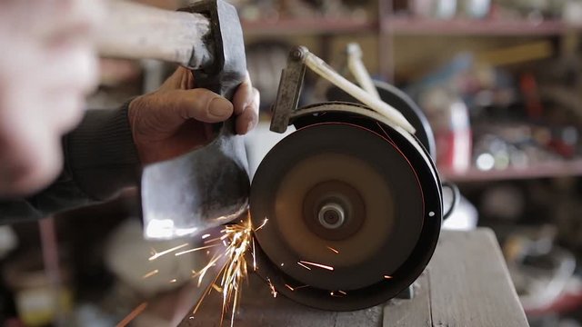 Old man sharpening his ax on a grinder in an workshop