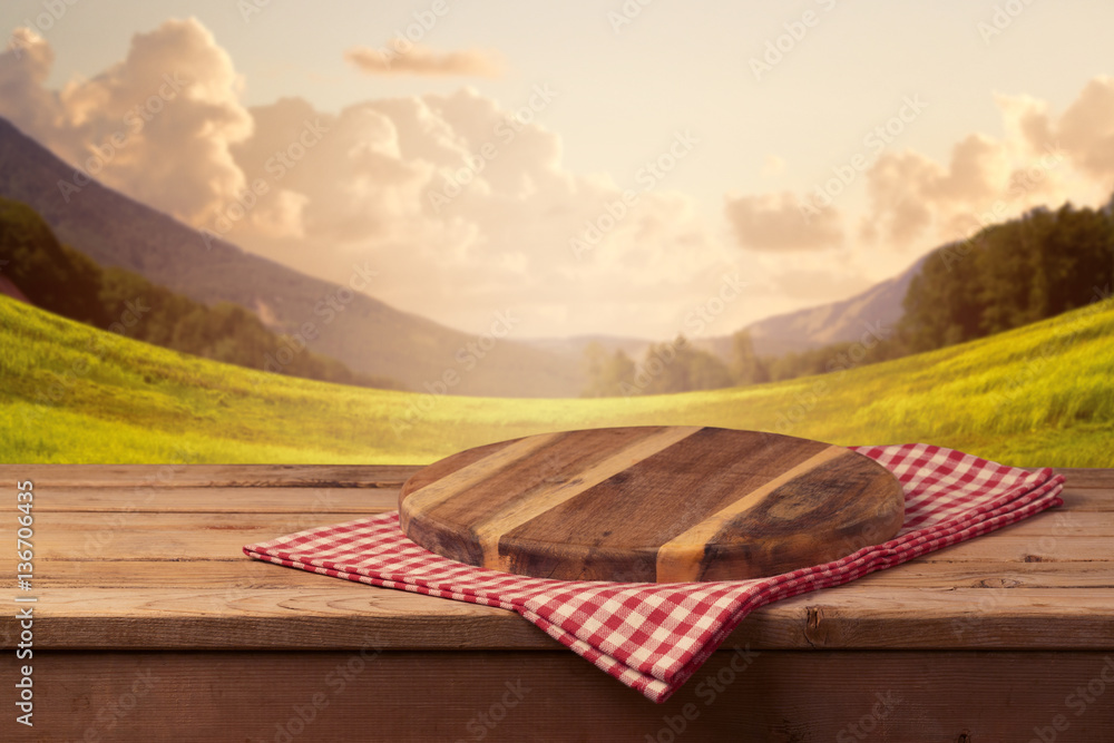 Wall mural wooden cutting board with checked tablecloth on table over beautiful landscape background