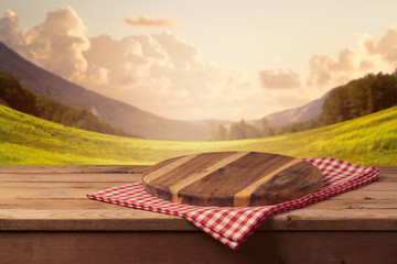 Wooden cutting board with checked tablecloth on table over beautiful landscape background