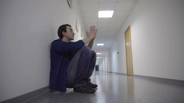 man squats in the long white corridor, leaning against the wall, praying, hoping and waits