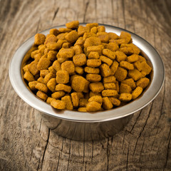 Bowl of dry kibble pets cat dog food on rough rustic wooden background. Top view, selective focus, square toned vignette mage.
