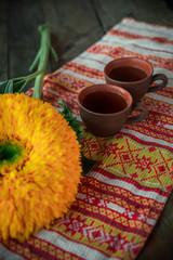On the table, on a towel with a national ornament clay cups of tea. Next to the flower of a sunflower.