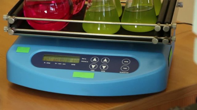 Laboratory workspace  panning. Camera moves down &ndash; from microscope oculars, rotating shaker with colorful glassware to green grass growing in glass container. Focus changes from shaker to grass.