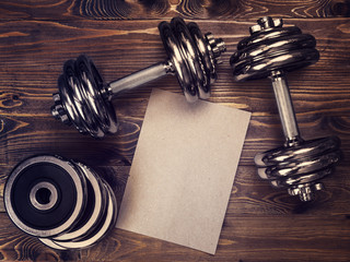 Toned image of metal dumbbells and a sheet of craft paper on a wooden background