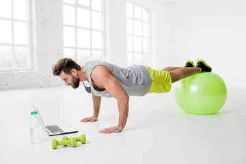 Man watching online trainig with laptop training on the fitball in the gym