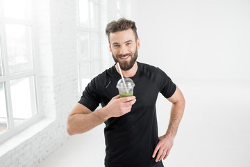 Handsome man in the black sportswear drinking green smoothie in the white gym interior