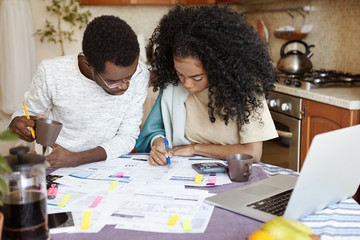 Naklejka premium Young African family having debt problems, not able to pay for gas and electricity, managing finances, sitting at kitchen table with papers, calculating bills, trying to cut their domestic expenses