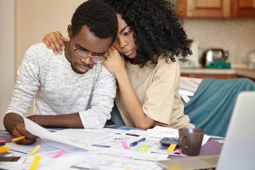 Unhappy young African couple facing financial stress: black man in spectacles holding notification informing that bank denied to prolong loan term, his beautiful wife embracing him supportively