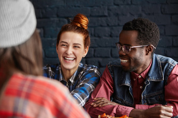 People, leisure, communication and friendship. Beautiful young woman with hair bun and her fashionable dark-skinned boyfriend laughing out loud while having lively conversation during breakfast