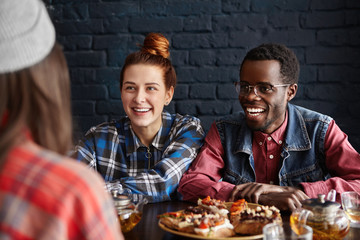 People, lifestyle, leisure and communication concept. Interracial group of three friends dining at restaurant, spending good time together, talking and sharing news, looking cheerful and happy