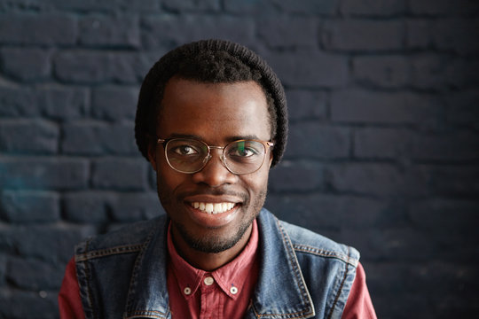 Indoor portrait of happy and cheerful young dark-skinned male student wearing trendy clothing looking at camera with joyful smile while having rest in modern cafe interior with black brick walls