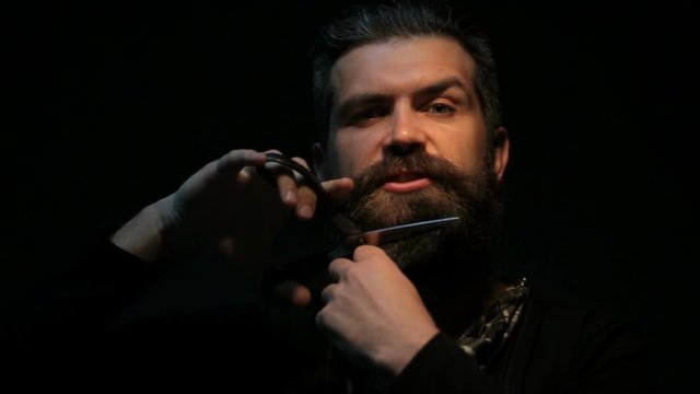 Bearded man with scissors cuts his beard. Barber makes a short beard himself. Attractive handsome man isolated on black background work with sharp scissors 