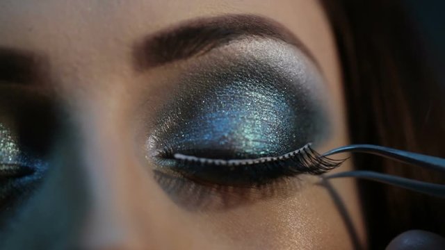 Eyelash extension, artificial eyelashes on the eyes with make-up, blue shadows. Makeup artist takes with forceps and stick artificial lashes on the woman's eye, close up, professional procedures